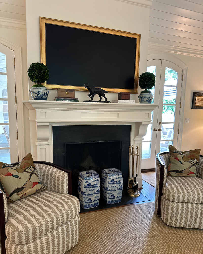 Timeless living room with a pair of swivel chairs, fireplace, chinoiserie garden stools and topiaries, and a gold frame TV